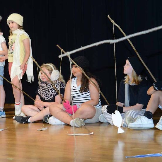 Eine Gruppe von Kindern ist auf der Bühne und hält Stöcke mit Schnüren und Fischpuppen. Sie sind in verschiedenen Kostümen gekleidet und scheinen eine Aufführung darzubieten.