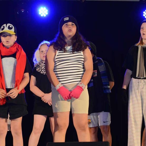 Eine Gruppe junger Kinder auf der Bühne, einige tragen Mützen und Handschuhe, unter blauen Bühnenlichtern.