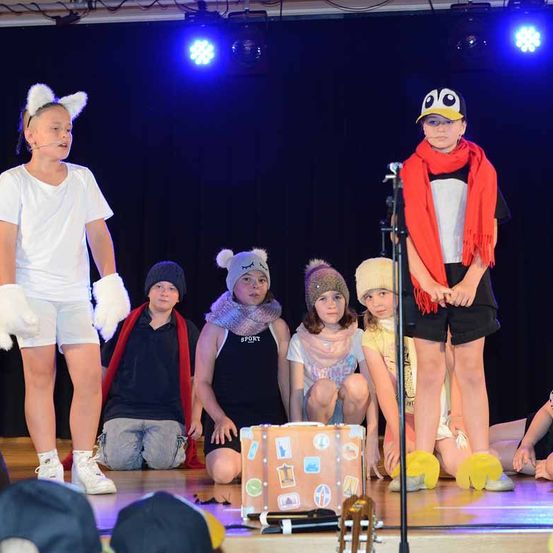 Kinder auf der Bühne in verschiedenen Kostümen. Eines trägt eine Pinguinmütze und -schal, ein anderes einen Katzenohren-Kopfschmuck. Ein Koffer mit Aufklebern steht zwischen ihnen, ein Mikrofon in der Nähe. Scheinwerfer beleuchten die Szene.