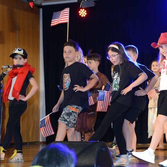 Eine Gruppe von Kindern in verschiedenen Kostümen tritt auf der Bühne auf. Einige von ihnen halten Flaggen. Vor einem Jungen mit rotem Schal steht ein Mikrofon.