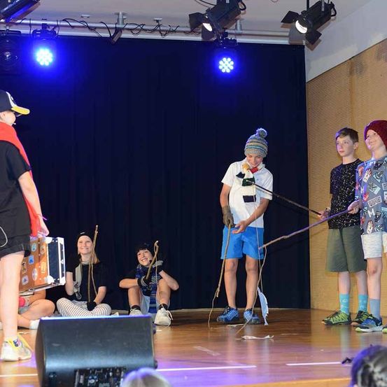 Mehrere Kinder stehen auf der Bühne, wahrscheinlich bei einer Aufführung. Ein Kind hält eine Trommel, und andere halten Stöcke. Scheinwerfer hängen an der Decke, und ein Lautsprecher steht auf dem Boden.