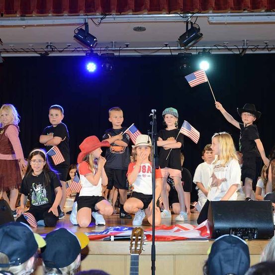Eine Gruppe von Kindern auf der Bühne führt einen patriotischen Akt mit amerikanischen Flaggen durch. Sie sind in verschiedenen thematischen Outfits gekleidet, einige knien und andere stehen. Scheinwerfer beleuchten die Bühne, und ein Lautsprecher ist rechts positioniert.