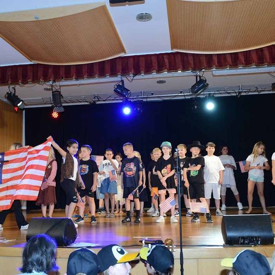 Eine Gruppe von Kindern steht auf der Bühne und hält amerikanische Flaggen in den Händen. Sie tragen verschiedene T-Shirts, Turnschuhe und Mützen. Vor ihnen steht ein Mikrofon, und es gibt Lautsprecher auf der Bühne. Scheinwerfer sind an der Decke angebracht.