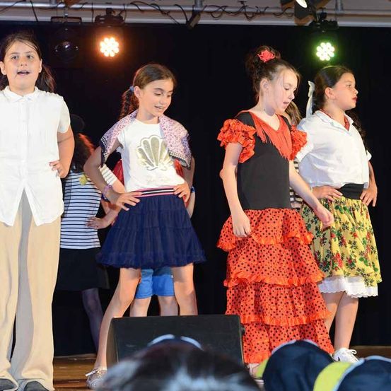 Eine Gruppe junger Mädchen in bunter Kleidung steht auf der Bühne und performt, mit zwei Scheinwerfern, die den Bereich beleuchten. Sie stehen in einer Reihe, möglicherweise singen und tanzen sie.