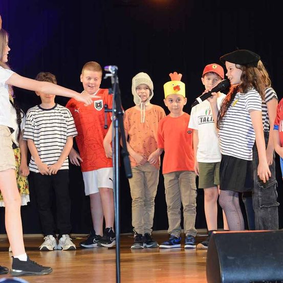 Eine Gruppe von Kindern steht auf der Bühne, einige in Kostümen. Ein Mädchen in einer schwarzen Beret hält ein Mikrofon, während andere in thematischen Outfits gekleidet sind.