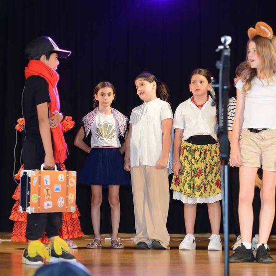 Eine Gruppe von Kindern steht auf der Bühne in einem Schulstück, einer hält einen Koffer und trägt einen Hut. Die Kinder sind in verschiedenen Kostümen.