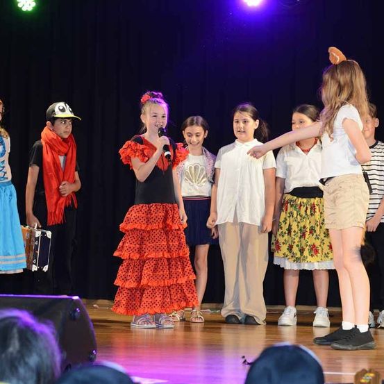 Eine Gruppe von Kindern in verschiedenen Kostümen tritt auf der Bühne auf. Ein Mädchen hält ein Mikrofon, während andere hinter ihr stehen. Der Hintergrund ist dunkel, und die Bühne wird von einem Scheinwerfer beleuchtet.
