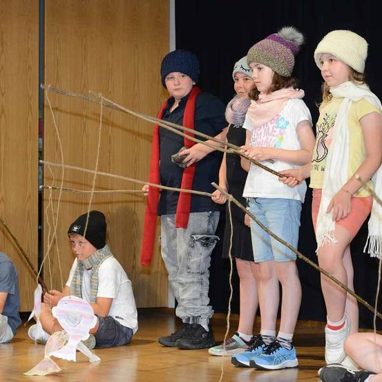 Eine Gruppe von Kindern hält Angelruten mit Fischpuppen an den Enden. Sie stehen auf einem Holzboden. Ein Junge sitzt auf dem Boden und hält eine Fischpuppe in den Händen.