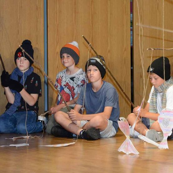 Fünf Jungen sitzen mit Angelruten auf dem Boden und tragen Winterkleidung. Sie scheinen an einer Aktivität mit Papierfischen beteiligt zu sein.