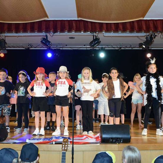 Kinder, die in USA-thematischer Kleidung gekleidet sind, treten auf einer Bühne auf, vor ihnen liegt eine US-Flagge und eine Menge schaut zu.