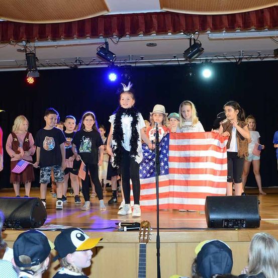 Eine Gruppe von Kindern steht auf der Bühne, einer hält eine amerikanische Flagge, während andere thematische Kostüme tragen und Flaggen halten. Die Bühne ist mit Lautsprechern und Scheinwerfern ausgestattet.