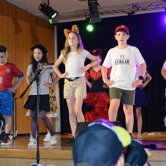 Kinder führen auf der Bühne auf, eines in einem roten Italien-Trikot, ein anderes in einem roten Slowenien-Trikot. Eine Mädchen mit Hasenohren und eine andere mit einem Beret stehen in der Nähe. Ein rotes Kleid und ein schwarzer Hut sind hinter ihnen zu sehen.