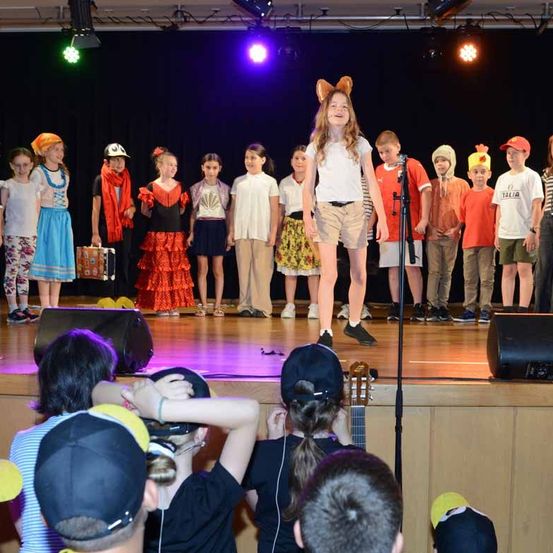 Eine Gruppe von Kindern tritt auf einer Bühne mit Mikrofon, Lautsprecher und bunten Lichtern auf. Ein Mädchen mit einer Katzen-Haarschleife steht in der Mitte. Das Publikum beobachtet sie.