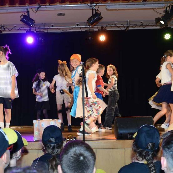 Kinder in verschiedenen Kostümen führen eine Aufführung auf der Bühne mit Scheinwerfern durch. Ein Kind hält ein Mikrofon. Zuschauer sehen von unten zu.