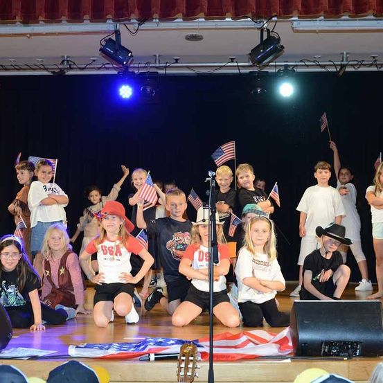 Eine Gruppe von Kindern auf der Bühne, einige stehend, andere sitzend, halten amerikanische Flaggen und führen eine patriotische Veranstaltung durch.