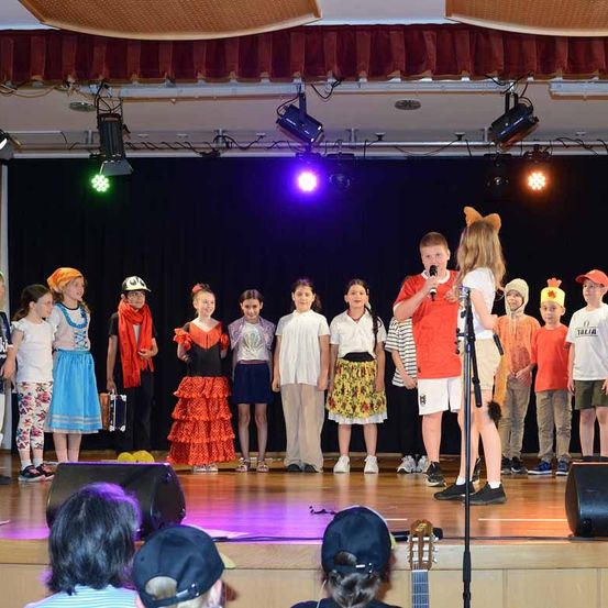 Eine Gruppe von Kindern auf der Bühne, einige halten Musikinstrumente, tragen verschiedene Kostüme und werden von Bühnenlichtern beleuchtet. Ein Kind hält ein Mikrofon.