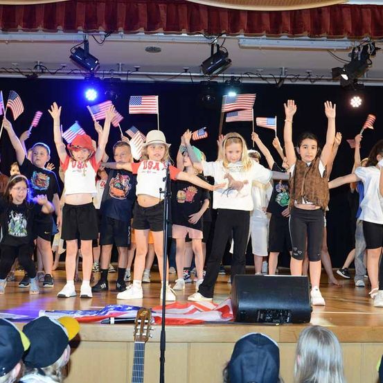 Eine Gruppe von Kindern und Erwachsenen tritt auf der Bühne auf und schwenkt amerikanische Flaggen. Ein Lautsprecher und eine Gitarre sind auf der Bühne platziert. Einige Leute schauen aus dem Publikum zu.