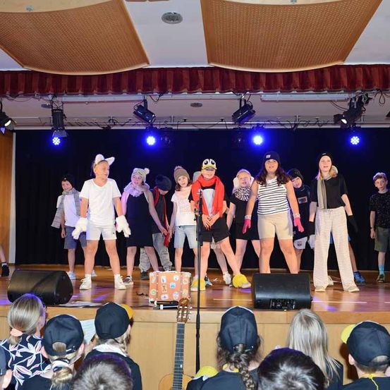 Eine Gruppe von Kindern führt mit verschiedenen Requisiten auf der Bühne auf, unter blauen Bühnenlichtern. Ein Kind hält ein Mikrofon. Ein Publikum sitzt vor ihnen.