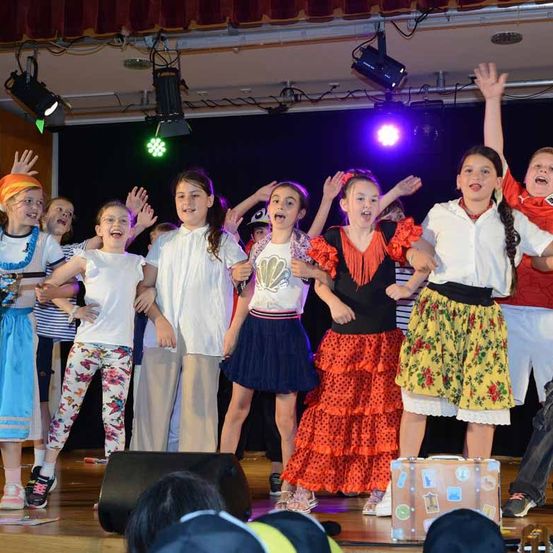 Eine Gruppe von Kindern auf der Bühne, wahrscheinlich in einem Schultheaterstück, mit bunten Kostümen und Lichtern.