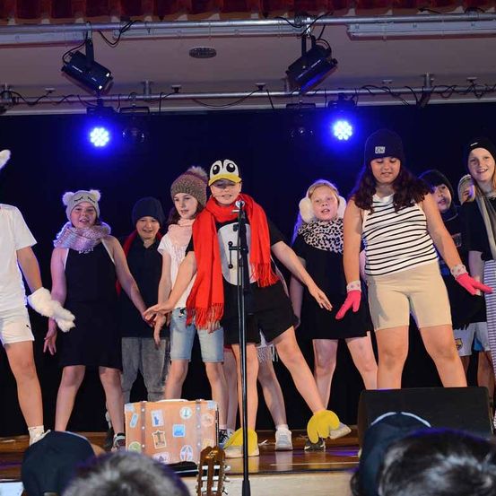 Eine Gruppe junger Darsteller auf der Bühne, einer hält ein Mikrofon. Sie tragen verschiedene Kostüme und Accessoires, mit blauen Bühnenlichtern im Hintergrund.