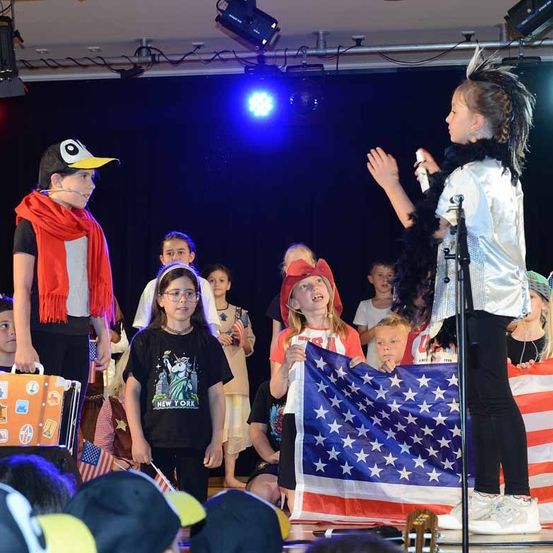 Eine Gruppe von Kindern steht auf der Bühne, einige halten amerikanische Flaggen und tragen verschiedene Kostüme. Ein Kind ist als Pinguin verkleidet und hält einen Koffer.