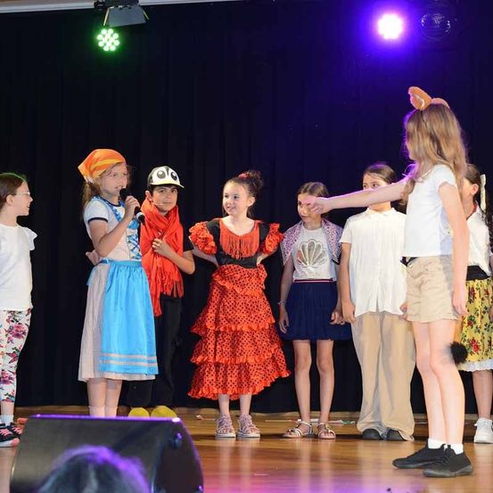 Kinder in verschiedenen Kostümen stehen auf der Bühne, einige halten Mikrofone. Ein Mädchen mit Hasenohren führt sie an. Die Bühne ist schwach beleuchtet mit lila Licht.