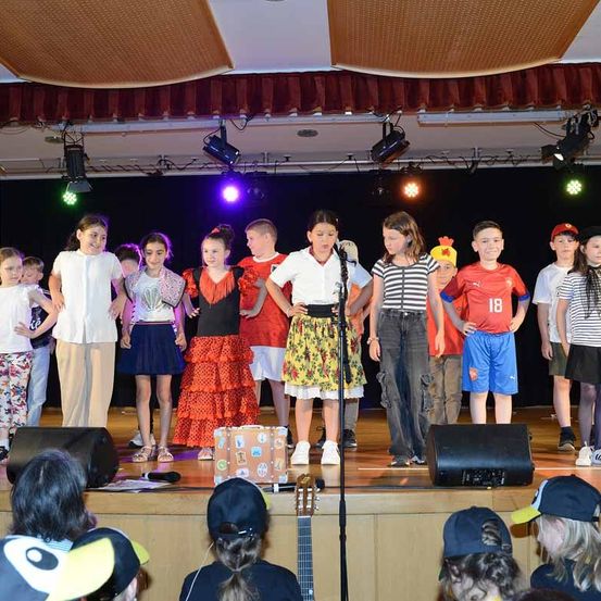 Eine Gruppe von Kindern tritt auf einer Bühne mit Mikrofonen auf, trägt verschiedene Kostüme und ist von Lautsprechern und einer Gitarre umgeben.