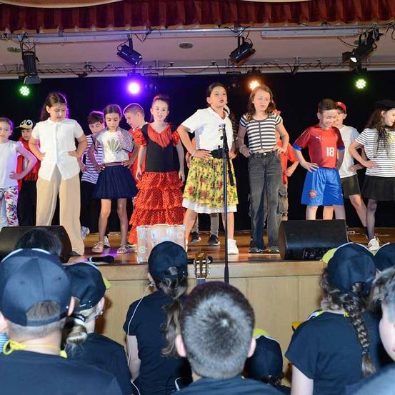 Eine Gruppe von Kindern tritt auf der Bühne auf, einige halten ein Mikrofon. Sie tragen verschiedene Kostüme, darunter ein rotes und schwarzes Kleid, ein weißes Hemd und ein gestreiftes Hemd. Die Bühne ist mit lila und grünen Lichtern beleuchtet.