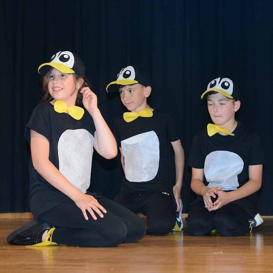 Drei Kinder in Pinguinkostümen sitzen auf dem Boden, wahrscheinlich auf der Bühne. Sie tragen schwarze T-Shirts mit Pinguindesign und gelbe Krawatten.