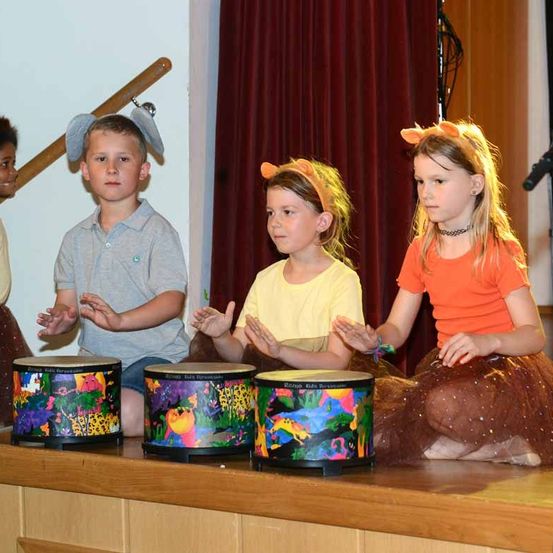 Drei Kinder mit Tierkopfbändern sitzen auf einer Bühne mit Trommeln vor ihnen. Der Junge trägt ein Elefantenkopfband, und die Mädchen tragen Löwen- und Tigerkopfbänder. Hinter ihnen ist ein roter Vorhang.