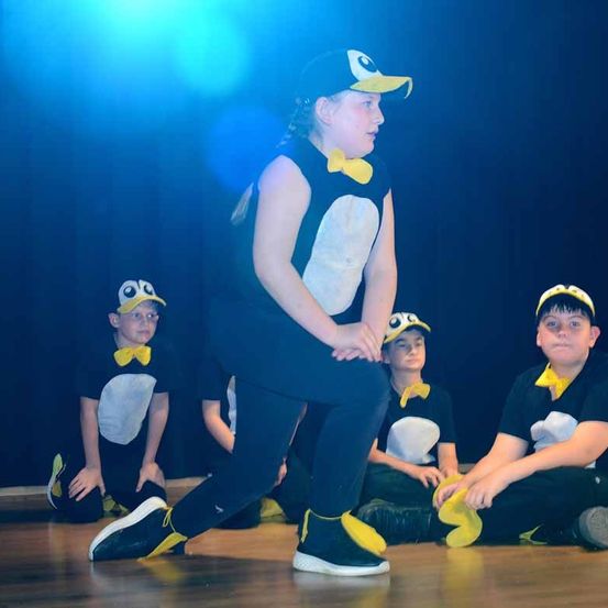 Eine Gruppe von Kindern, die als Pinguine verkleidet sind, tritt auf der Bühne auf, wobei ein Kind steht und andere dahinter sitzen. Sie stehen unter einem blauen Licht.