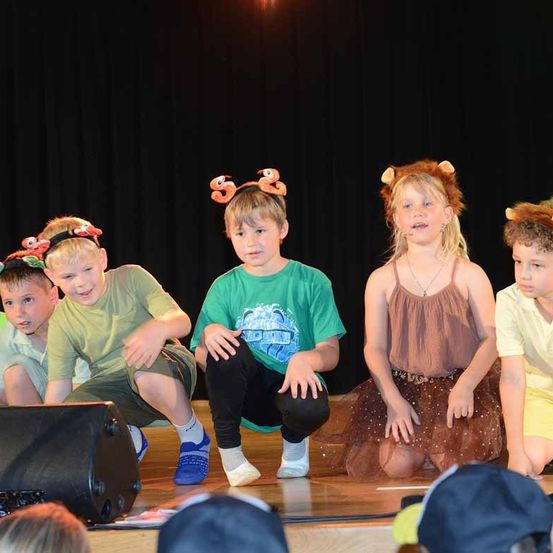 Eine Gruppe von Kindern auf der Bühne mit verschiedenen Tierkopfbändern, eines mit einem Löwenkopfband, ein anderes mit einem Rentierkopfband und ein Mädchen in einem braunen Kleid.