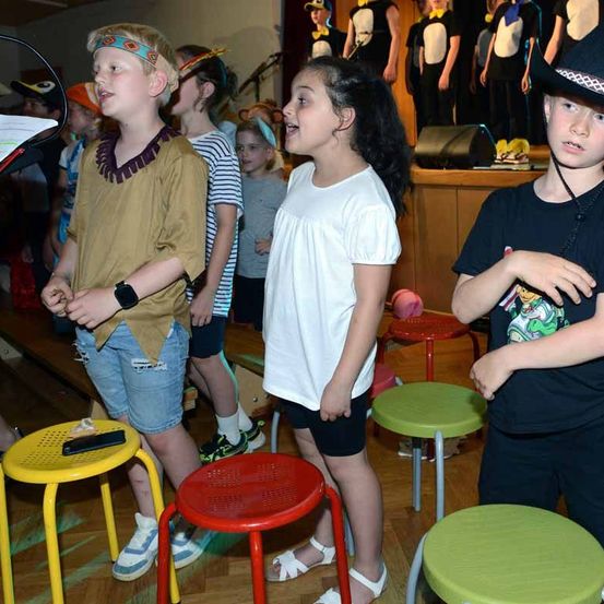 Mehrere Kinder in Freizeitkleidung stehen auf Hockern, möglicherweise bei einer Schulaufführung. Dahinter stehen andere Kinder, einige sitzen. Einige tragen Kostüme.