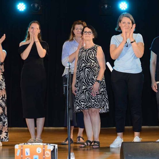Eine Gruppe von Frauen steht auf der Bühne, klatscht und lächelt, wobei eine Frau ein Mikrofon hält. Hinter ihnen steht ein Lautsprecher und ein Koffer mit Aufklebern.
