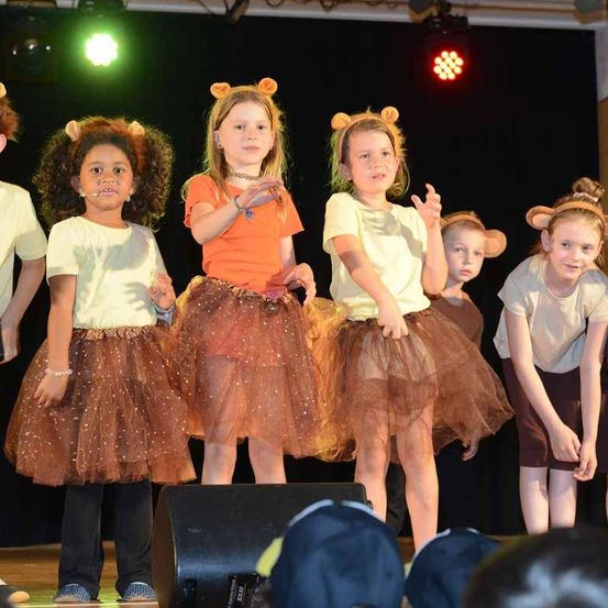 Mehrere Kinder stehen auf der Bühne und spielen, alle in Kostümen gekleidet. Eines der Mädchen trägt ein orangefarbenes T-Shirt. Sie nehmen wahrscheinlich an einem Schultheaterstück oder einer Veranstaltung teil.