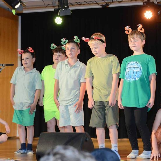 Eine Gruppe von Kindern mit Wurmkopfbändern auf der Bühne. Der Junge in Grün mit 'HOPE' auf seinem Shirt schaut nach unten. Die Bühne hat ein Mikrofon und einen Lautsprecher. Scheinwerfer sind an der Decke.