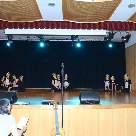 Eine Gruppe von Kindern in Pinguinkostümen tritt auf einer Bühne mit Scheinwerfern auf, während eine Lehrerin von der Seite zusieht.