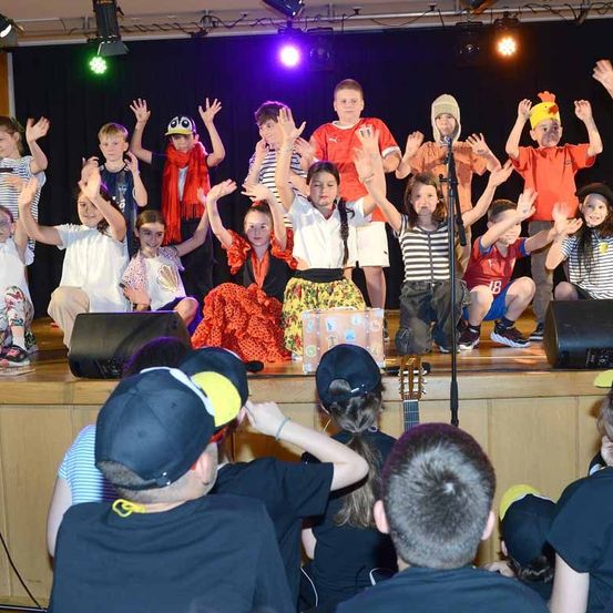 Eine Gruppe von Kindern tritt auf einer Bühne auf, hebt die Hände und trägt verschiedene Kostüme. Einige haben Hüte, und andere haben Schals. Ein Mikrofon steht vor ihnen, und Lautsprecher sind auf der Bühne platziert. Das Publikum beobachtet von vorne.