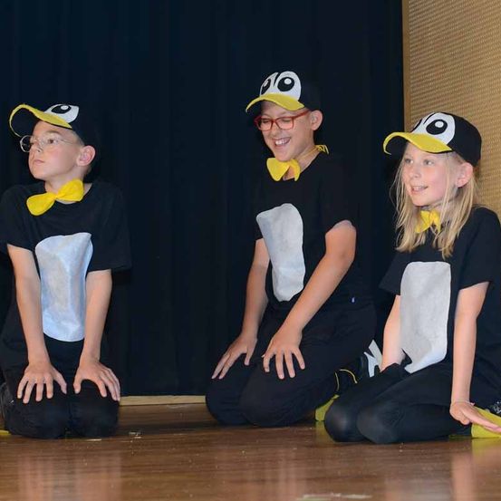 Drei Kinder in Pinguinkostümen sitzen auf einer Bühne, lächeln und haben einen dunklen Hintergrund hinter sich.