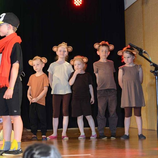 Eine Gruppe von Kindern mit Tierohren-Kopfbändern steht auf der Bühne. Sie sind in verschiedenen farbigen Kleidern gekleidet. Ein Mikrofon auf einem Ständer ist auf der Bühne vorhanden.