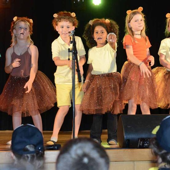 Eine Gruppe von Kindern in Löwenkostümen tritt auf der Bühne auf, mit einem Mikrofon vor ihnen. Einige tragen braune Tüllröcke und andere verschiedene Oberteile. Ein Lautsprecher steht rechts auf der Bühne.