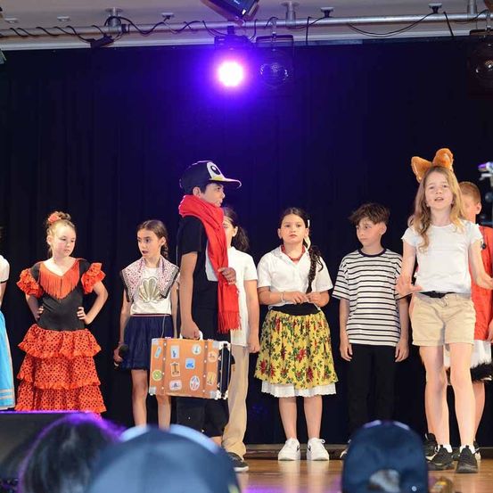 Eine Gruppe von Kindern steht auf der Bühne in Kostümen. Ein Kind hält einen Koffer, und andere tragen Hüte. Sie stehen vor einem blauen Vorhang, und es gibt Lichter über ihnen.