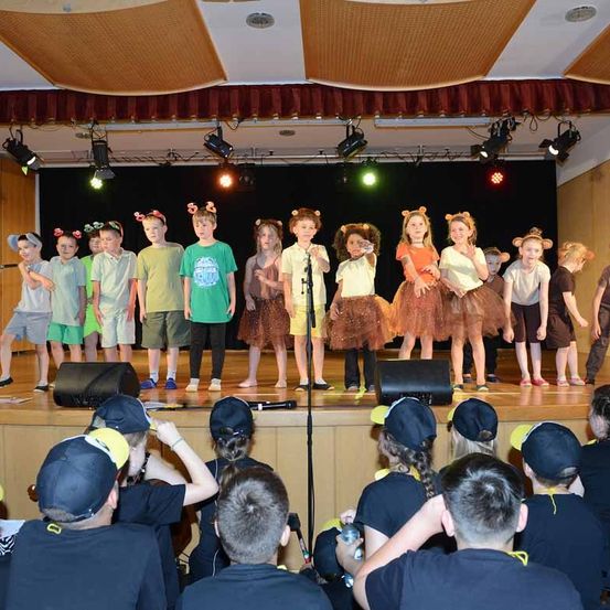 Eine Gruppe von Kindern in Kostümen und Tierohren führt auf einer Bühne mit Mikrofonen und Lautsprechern auf, während ein Publikum sie beobachtet.