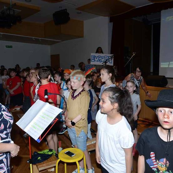 Kinder versammeln sich in einem Raum für eine Veranstaltung, einige tragen Hüte. Eine Person steht vor ihnen mit einem Mikrofon. Eine Erwachsene hält ein Schild mit dem Wort 'Europa'.