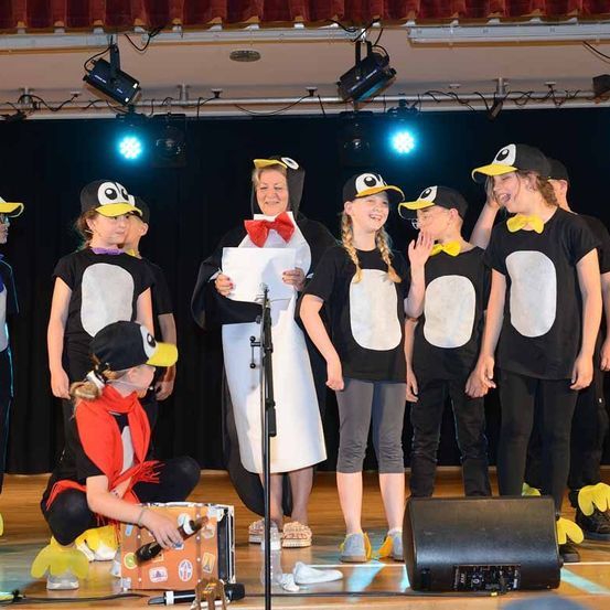 Eine Gruppe von Kindern in Pinguinkostümen, die auf der Bühne auftreten, einer hält ein Mikrofon und ein Papier, andere stehen in einer Reihe mit einem Lautsprecher und einer Frau mit einer Schleife.
