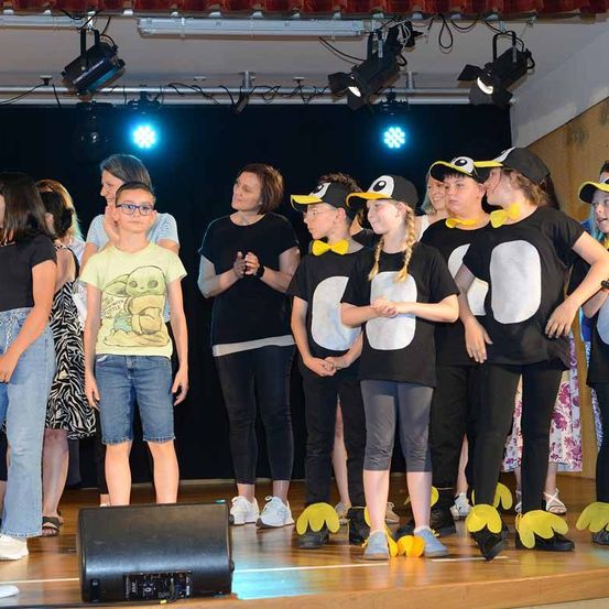 Eine Gruppe von Kindern und Erwachsenen steht auf der Bühne in Pinguinkostümen. Zwei Frauen hinter ihnen klatschen. Ein Lautsprecher befindet sich auf der linken Seite.