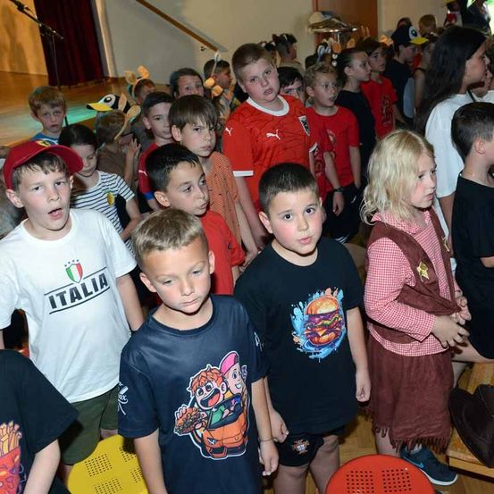 Eine Gruppe von Kindern, einige in Kostümen, stehen in einem Saal, wahrscheinlich für eine Schulveranstaltung. Einige tragen T-Shirts mit gedrucktem Text.