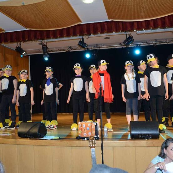 Eine Gruppe von Kindern in Pinguinkostümen tritt auf der Bühne auf, mit zwei schwarzen Lautsprechern, einer Gitarre und einer Person in der vorderen Reihe. Scheinwerfer sind an der Decke montiert.