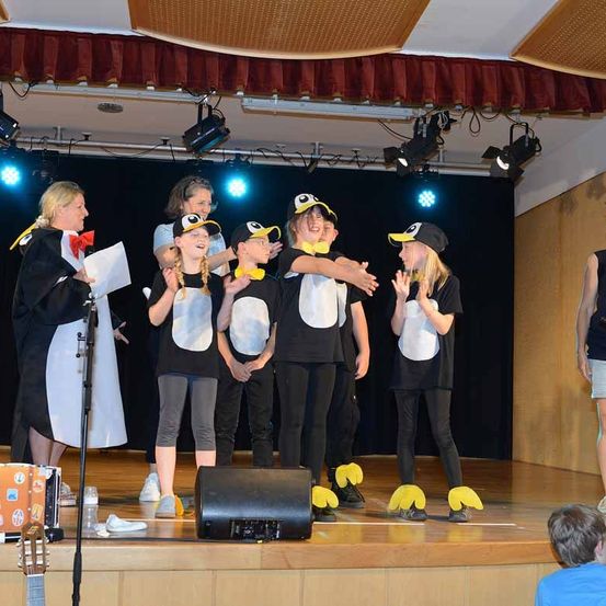 Eine Gruppe von Kindern in Pinguinkostümen tritt mit einer Frau und einem Lautsprecher auf der Bühne auf. Das Publikum beobachtet von unten.