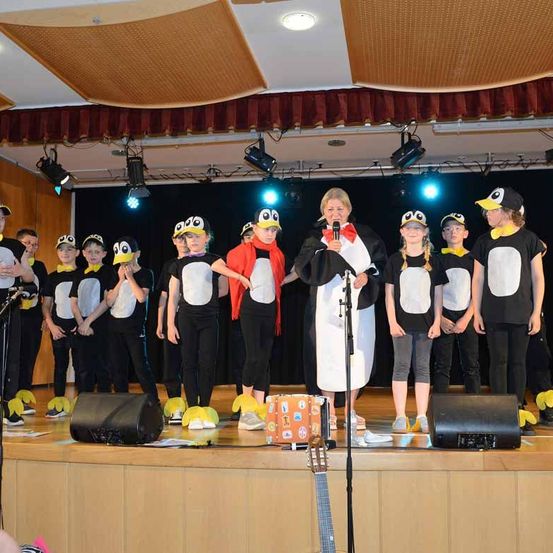 Eine Frau mit einem Mikrofon steht auf der Bühne mit einer Gruppe von Kindern in Pinguinkostümen. Hinter ihnen sind Scheinwerfer an. Vor der Frau steht eine Gitarre.